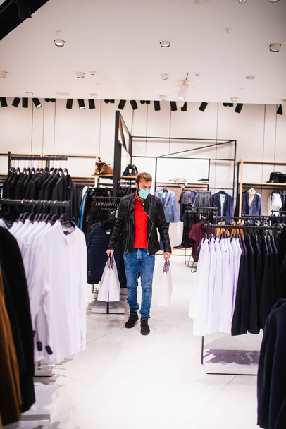 Un giovane uomo caucasico urbano con una maschera medica e borse esamina un armadio in una boutique. Shopping nel centro commerciale durante l'epidemia di COVID - 19 coronavirus - Foto, immagini