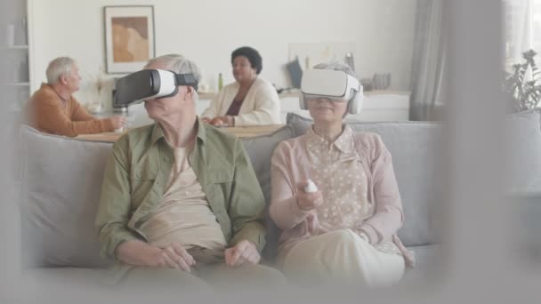 Mittellange grauhaarige Männer und Frauen, die Virtual-Reality-Headsets tragen, auf der Couch im Wohnzimmer sitzen, die Köpfe seitlich bewegen, rauf und runter, reden, lächeln - Filmmaterial, Video