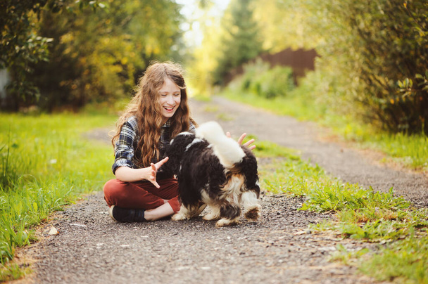 happy kid girl walking with her cavalier king charles spaniel dog on summer country road. Training her puppy and having fun.  - Zdjęcie, obraz