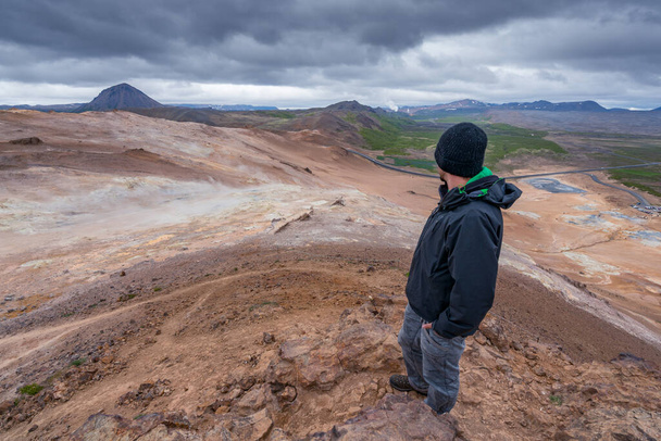 Namafjall 'ın tepesinden volkanik İzlanda manzarasına bakan bir adam. Gökyüzünde dramatik bulutlar. Uzaktaki yol karadan esen rüzgarlar gibi. Hver, çamur sahası kaynıyor.. - Fotoğraf, Görsel
