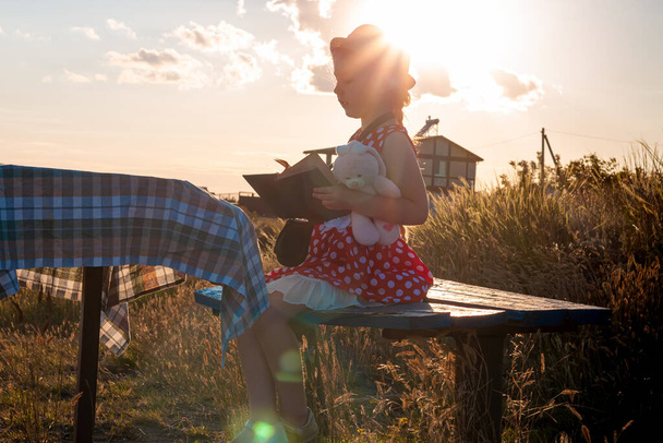 Ein kleines Mädchen mit Hut und gepunktetem Kleid sitzt auf einer Bank und liest ein Buch. Kind betrachtet die Heilige Bibel in Händen und betet auf Meereslandschaft Hintergrund. Freundschaft Frieden Religion Glaube Hoffnung Konzept. - Foto, Bild