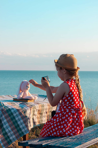 Hasır şapkalı küçük fotoğrafçı çocuk ve klasik bankta kırmızı benekli elbise deniz manzarasında yumuşak pembe tavşan oyuncağının fotoğrafını çekiyor. Kız elindeki kameraya bakıyor. Arkadaşlık eğlence seyahati. - Fotoğraf, Görsel