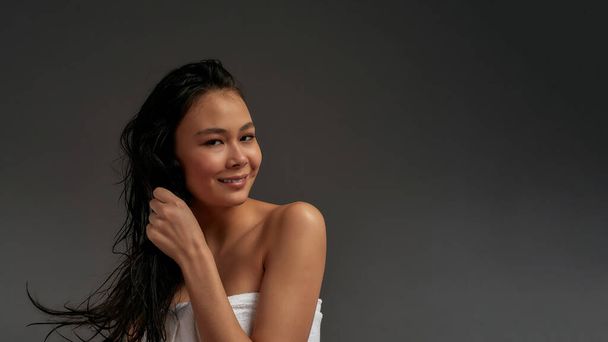 Nuori kaunis aasialainen puoli alasti nainen pörrötti hiuksiaan yhdellä kädellä kylpypyyhkeessä katsoo kameraan - Valokuva, kuva