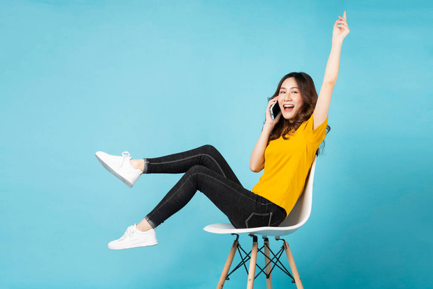 Νεαρό κορίτσι από την Ασία κάθεται στην καρέκλα χρησιμοποιώντας το τηλέφωνο με χαρούμενη έκφραση στο παρασκήνιο - Φωτογραφία, εικόνα