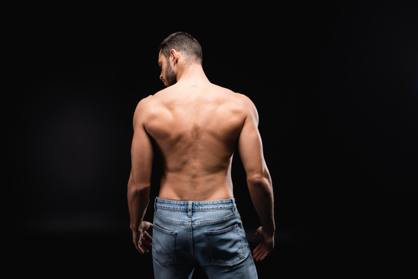 vista posterior de hombre sin camisa, musculoso en jeans sobre fondo negro - Foto, Imagen
