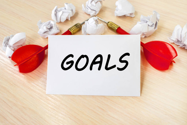 The word goal is written on a white sheet of paper that stands on a wooden table against a background of two red darts and crumpled sheets of paper. - Photo, image
