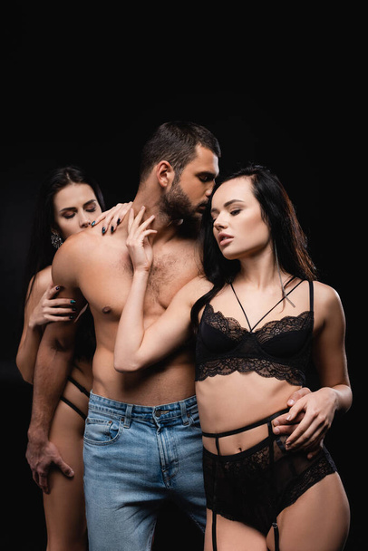 passionate women in lingerie seducing shirtless man isolated on black - Photo, Image