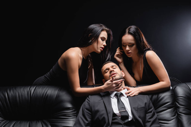 passionate, brunette women seducing businessman sitting on leather couch on black - Foto, Imagem