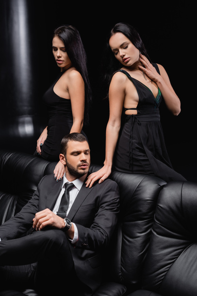 morena mujeres en elegantes vestidos tocando hombros de hombre de negocios sentado en el sofá de cuero en negro - Foto, Imagen