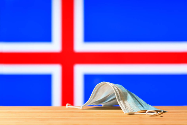 Una maschera medica si trova sul tavolo sullo sfondo della bandiera islandese. Il concetto di un regime di maschera obbligatorio per i residenti del paese e i turisti in Islanda durante una pandemia. - Foto, immagini