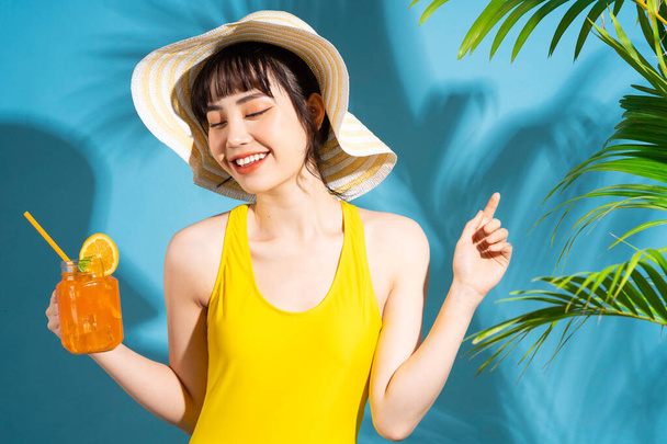 Schöne Asiatin in gelbem Overall auf blauem Hintergrund und trinkt Saft, Sommerkonzept - Foto, Bild