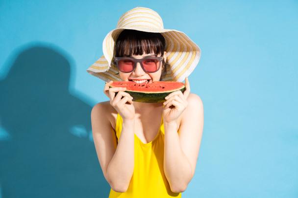Beautiful Asian woman wearing yellow jumpsuit on blue background and eating tropical fruits, summer concept - Photo, Image
