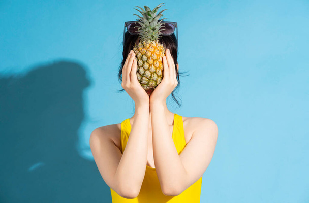 Beautiful Asian woman wearing yellow jumpsuit on blue background and eating tropical fruits, summer concept - 写真・画像