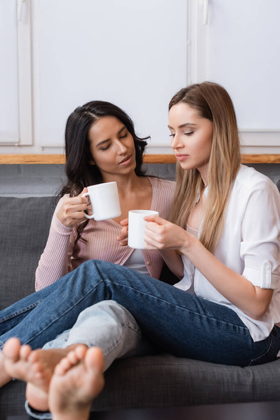 Freundinnen halten Tassen Tee, während sie auf dem Sofa im Wohnzimmer sitzen - Foto, Bild