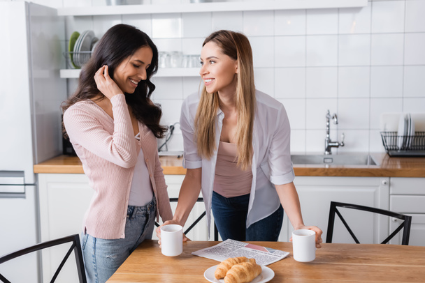 happy woman looking at shy girlfriend in kitchen  - Photo, Image