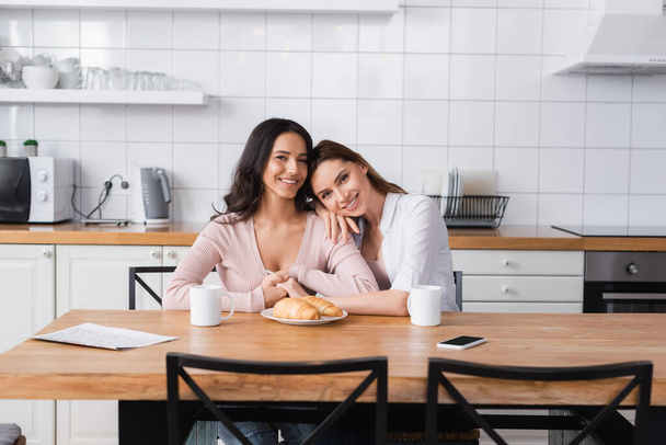 happy lesbian couple hugging near breakfast and smartphone on kitchen table  - Foto, Imagen