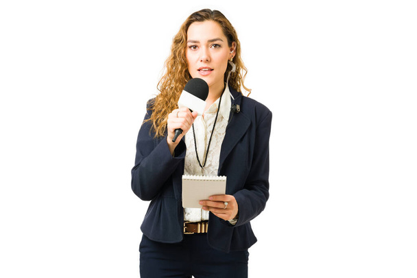 Retrato de un hermoso periodista sosteniendo un cuaderno y un micrófono mientras habla de noticias de última hora durante un programa de televisión en vivo - Foto, Imagen