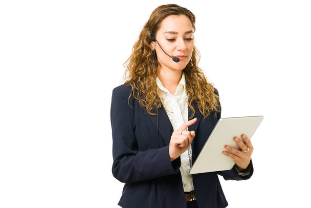 Mujer profesional ocupada usando una tableta e información de lectura mientras habla con un cliente con un auricular. Mujer joven hispana haciendo una venta en el trabajo - Foto, Imagen