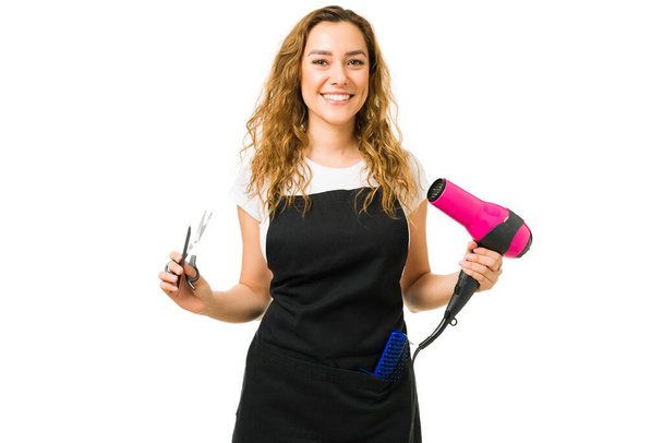 Happy attractive young woman smiling while making eye contact and holding a blow dryer with some scissors. Hair stylist working at a beauty salon - Photo, Image