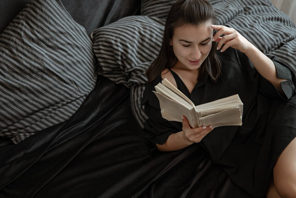 Aantrekkelijke jonge vrouw in pyjama is het lezen van een boek terwijl liggend in bed. - Foto, afbeelding
