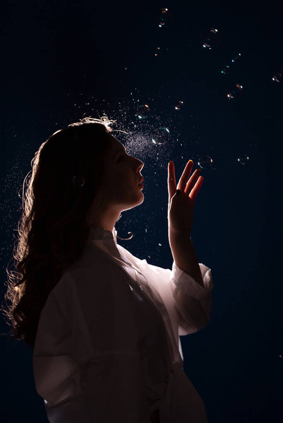 young woman in the dark in front of a bright light in a spray of soap bubbles and puffs of smoke - 写真・画像