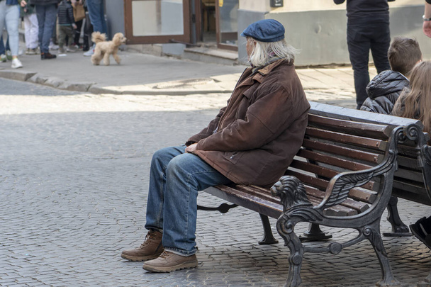 Уличный портрет пожилого человека 60-69 лет, сидящего на скамейке в центре города на пешеходной улице. Возможно, он безработный пенсионер или бывший военный, бездомный или больной.. - Фото, изображение