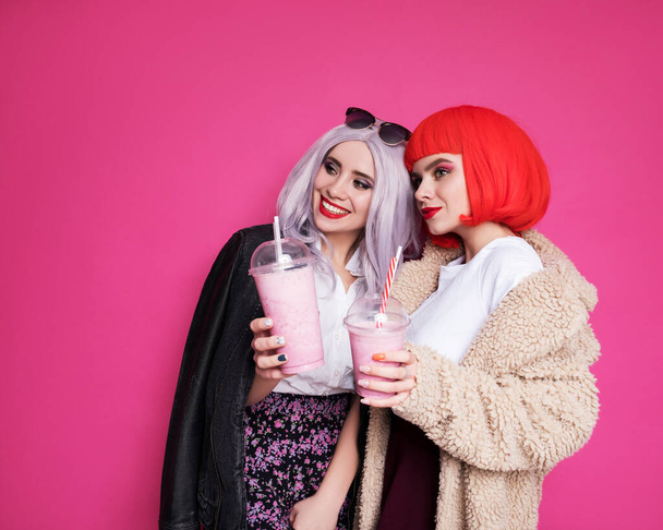 Dos amigas lindas mujer que parecen adolescentes de pie sobre un fondo rosa. Llevan pelucas de colores y elegantes trajes casuales y comer pizza con batidos. Felices retratos emocionales - Foto, imagen