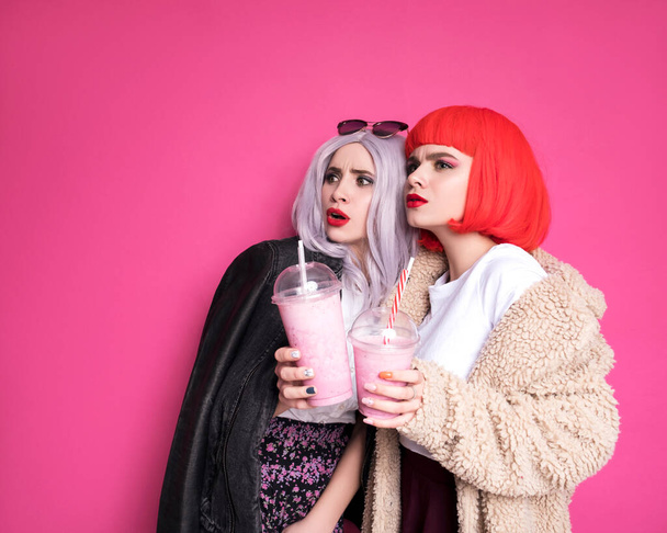 Twee leuke vriendinnen die eruit zien als tieners die op een roze achtergrond staan. Ze dragen kleurrijke pruiken en stijlvolle casual outfits en eten pizza met milkshakes. Gelukkige emotionele portretten - Foto, afbeelding