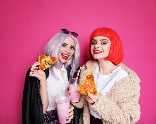 Dos amigas lindas mujer que parecen adolescentes de pie sobre un fondo rosa. Llevan pelucas de colores y elegantes trajes casuales y comer pizza con batidos. Felices retratos emocionales - Foto, Imagen