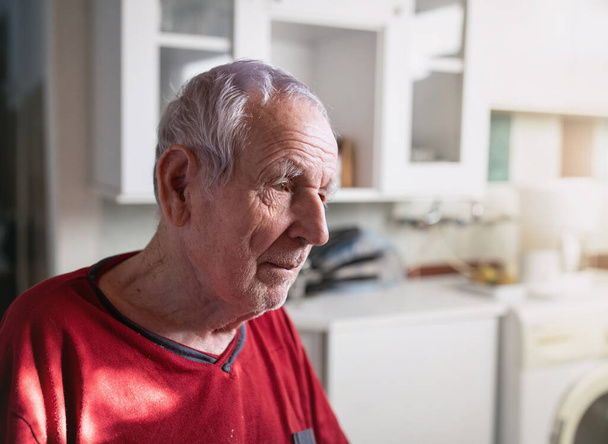 A 80-as évei végén járó ráncos bőrű öregember portréja otthon ül a konyhában. - Fotó, kép