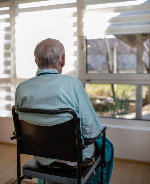 Вид ззаду старшого чоловіка в піжамі, сидячи в інвалідному кріслі і дивлячись через вікно в лікарні
 - Фото, зображення