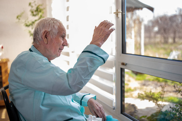 Senior man sitting in wheelchair, looking through big window and waving hand for greeting. Social distancing concept - Foto, afbeelding