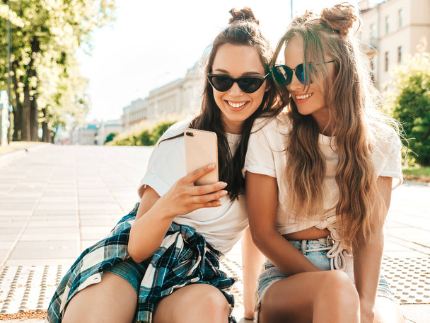 Portrait of two young beautiful smiling hipster women in trendy summer white t-shirt clothes.Sexy carefree women posing on street background. Positive models looking at cellphone screen. Using apps - 写真・画像