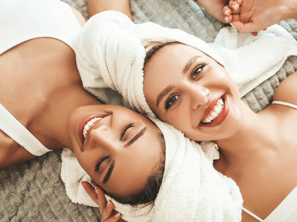 Two young beautiful smiling women in white bathrobes and towels on head.Sexy carefree models lying on bed in posh apartment. They doing beauty treatments at home in towels on heads.After face massage - Photo, image