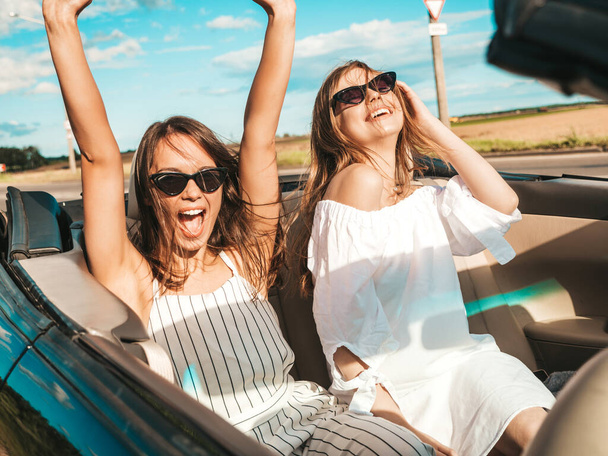Portrait of two young beautiful and smiling hipster female in convertible car. Sexy carefree women driving cabriolet. Positive models riding and having fun in sunglasses outdoors.Enjoying summer days - Фото, изображение