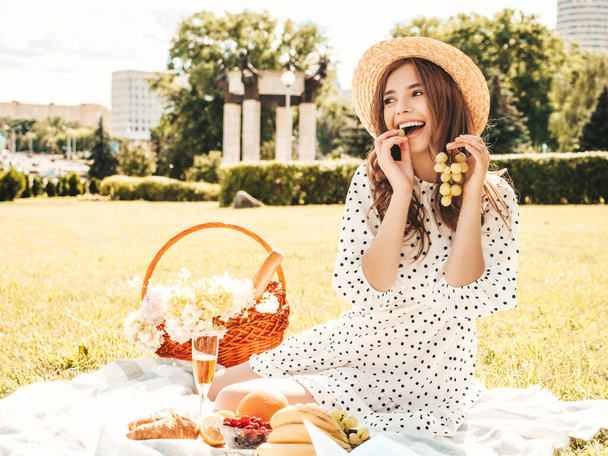 Young beautiful hipster woman in trendy summer jeans, pink T-shirt and hat. Carefree woman making picnic outside. Positive model sitting on plaid on grass, eating fruits and cheese, reading book - Zdjęcie, obraz