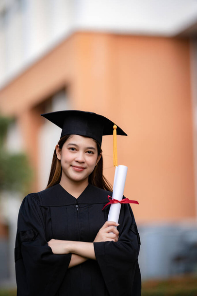 A young happy Asian woman university graduate in graduation gown and mortarboard holds a degree certificate celebrates education achievement in the university campus.  Education stock photo - Foto, imagen