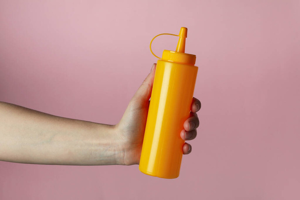 Female hand hold yellow sauce bottle on pink background - Foto, imagen