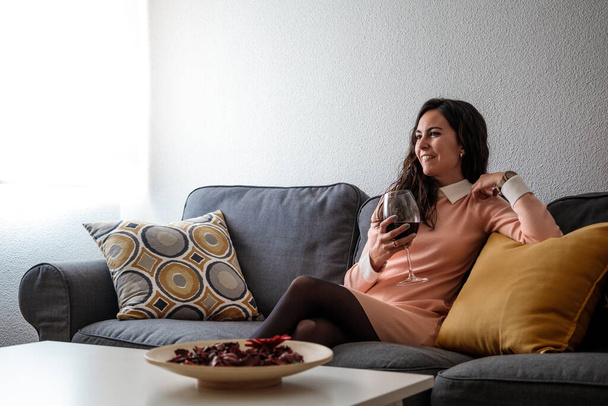 Ελκυστική νεαρή γυναίκα χαλαρώνει στον καναπέ της στο σπίτι με ένα ποτήρι λευκό κρασί. - Φωτογραφία, εικόνα
