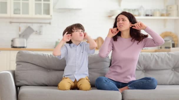 Fruncir el ceño mamá y el hijo se sientan en el sofá con los ojos cerrados y cubren los oídos de la música ruidosa o sonidos de lucha - Imágenes, Vídeo