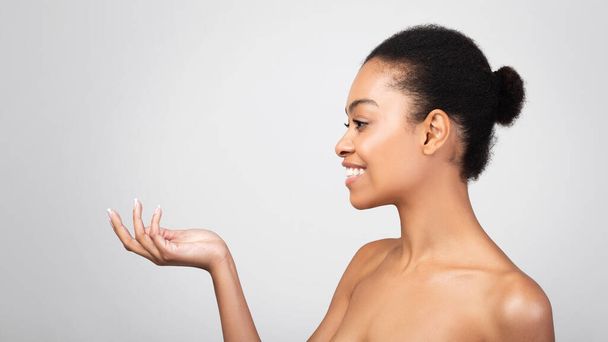 Mujer negra sosteniendo invisibles cosméticos tarro publicidad producto, fondo gris - Foto, Imagen