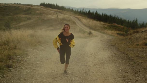 Κορίτσι τρέχει γρήγορα στο βρώμικο δρόμο. Χαμογελαστή αθλητική προπόνηση γυναικών στα βουνά - Φωτογραφία, εικόνα