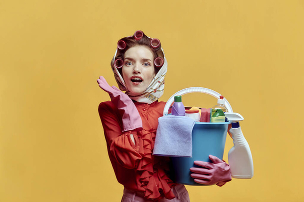A young surprised female cleaner holds a plastic bucket with cleaning tools. - 写真・画像
