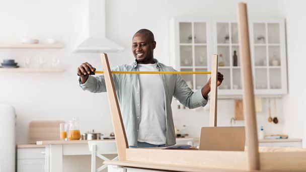 Emocionado hombre negro que mide muebles con ruleta, montando una nueva mesa en el interior de la cocina - Foto, Imagen