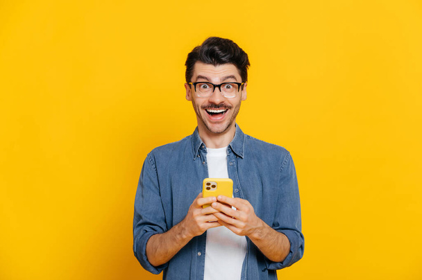 Happy komea hämmästynyt tyylikäs kaveri silmälasit, älypuhelimella, chattailuun verkossa, kirjallisesti viesti, selailu internet, sosiaalinen media, näyttää yllättynyt kamera, seisoo eristetty oranssi - Valokuva, kuva