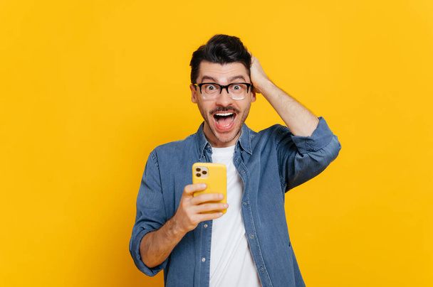 Amazed shocked positive caucasian guy with glasses, uses smartphone, browsing Internet, social networks, get unexpected news, holding his head with his hand, stands on isolated orange background - Photo, Image