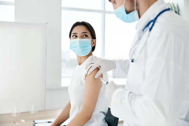 Tıbbi önlüklü bir doktor, tıbbi maskeli bir hastanın omzuna aşı enjekte ediyor.  - Fotoğraf, Görsel