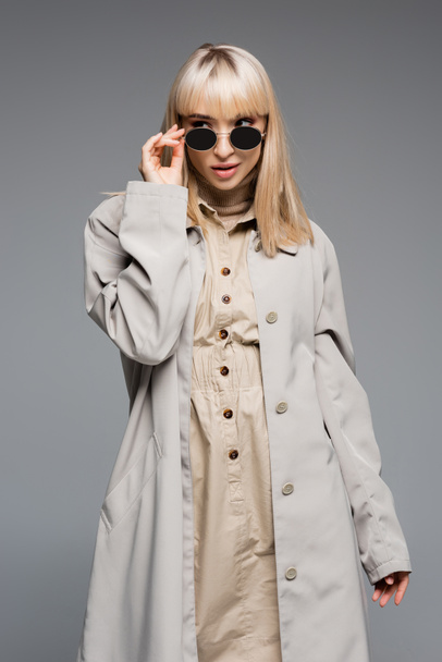 trendy young woman in trench coat adjusting sunglasses isolated on grey - Photo, image