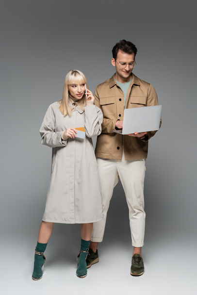 young woman in shirt dress talking on smartphone and holding credit cards near stylish man using laptop on grey - Foto, Bild