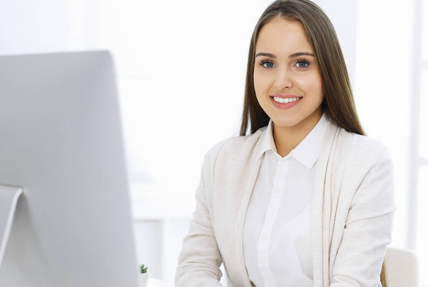 Young cheerful woman sitting at the desk with computer and looking at camera in white colored office. Looks like student girl or business lady. Headshot - 写真・画像